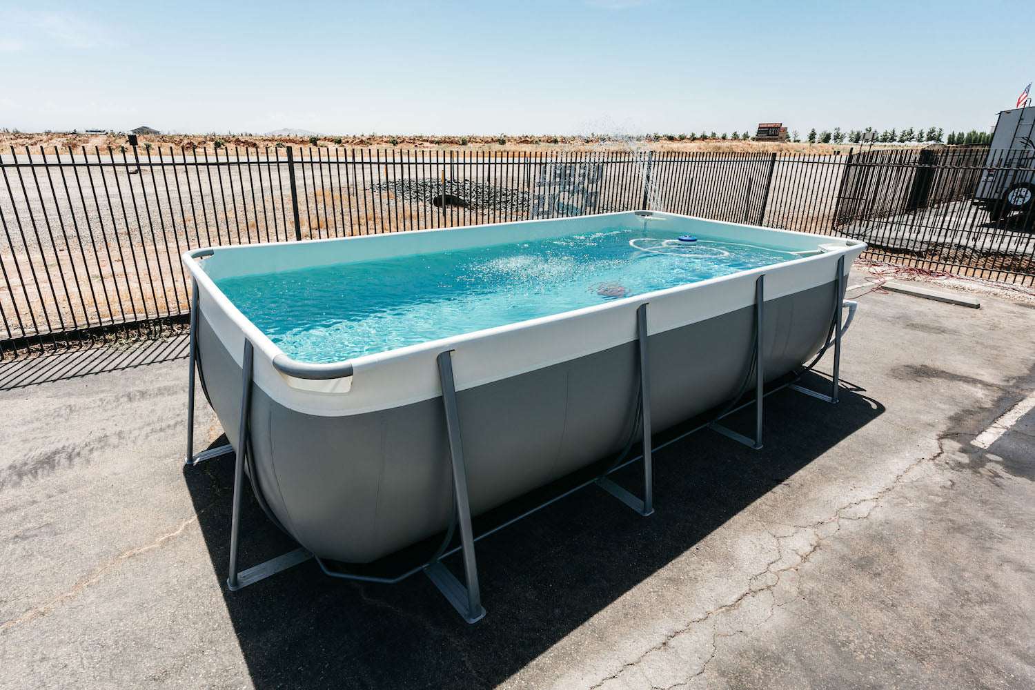The Kona Pools Rectangle Pool stands filled with water on asphalt near a metal fence, with dry grass and trees in the background under a clear blue sky.