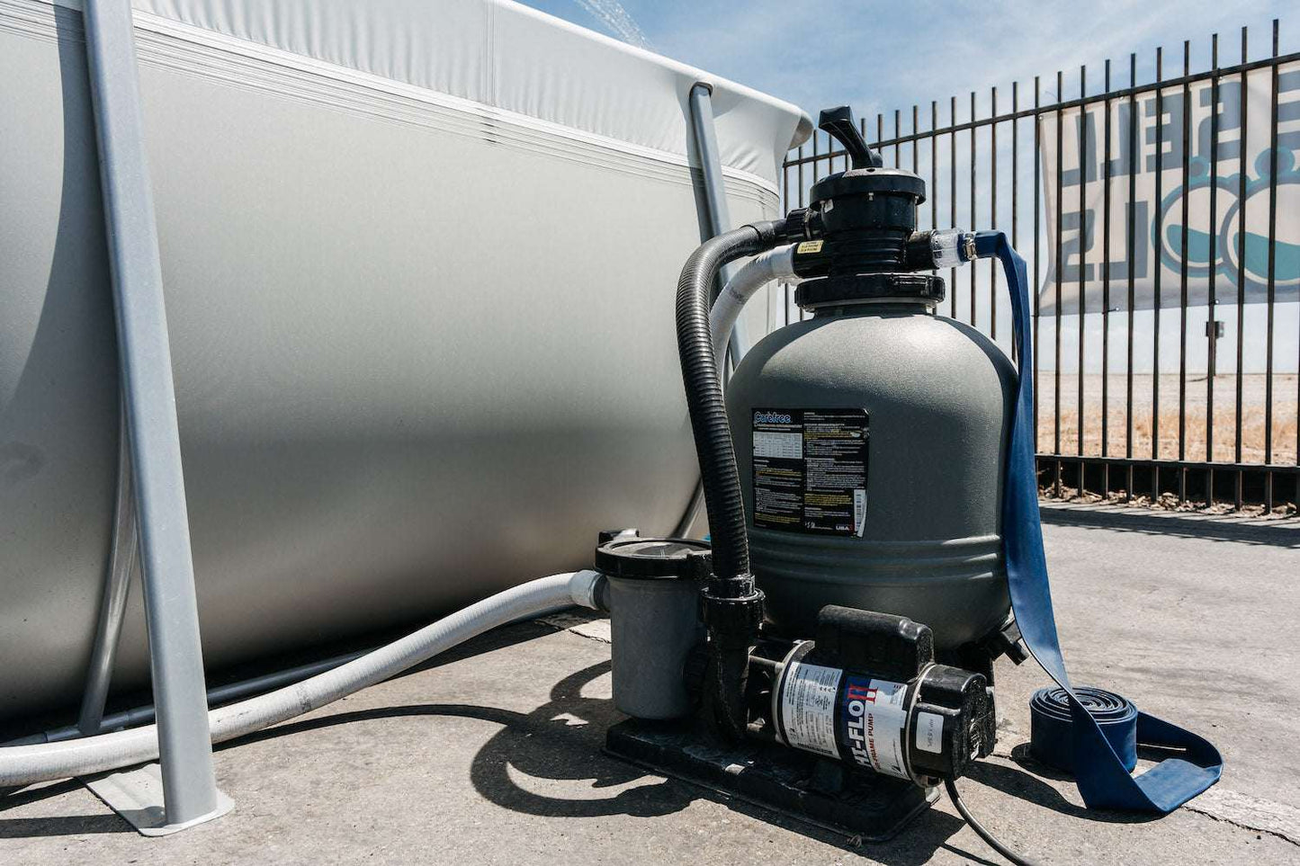 A My Store Waterway Sand Filter with hoses is connected to a Kona Above Ground Pool outdoors near a metal fence on a sunny day.