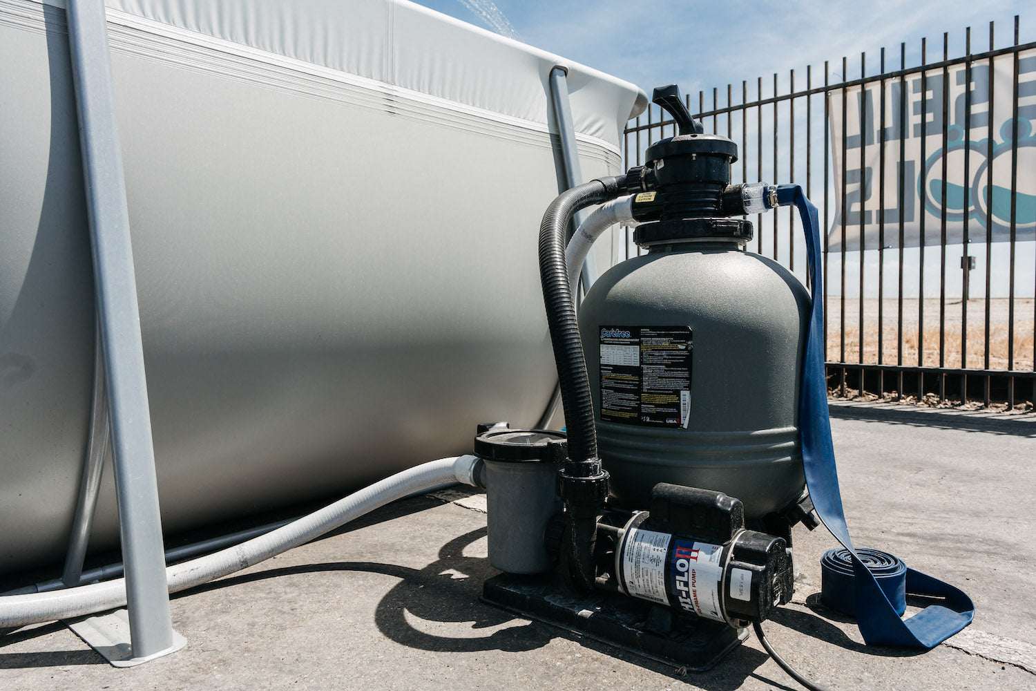 A My Store Waterway Sand Filter with hoses is connected to a Kona Above Ground Pool outdoors near a metal fence on a sunny day.