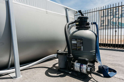 A My Store Waterway Sand Filter with hoses is connected to a Kona Above Ground Pool outdoors near a metal fence on a sunny day.