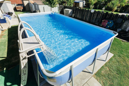 An above-ground backyard pool with clear blue water and a metal frame features the My Store Confer A Frame Pool Ladder. The pool is surrounded by a fence, grass, nearby sheds, and trees.