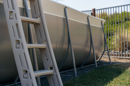A close-up of an above ground pool with metal supports features the My Store Confer A Frame Pool Ladder in beige, with a metal fence and greenery in the background.
