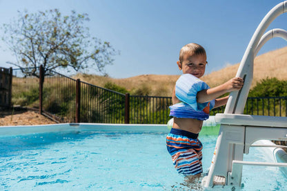 A young boy in a striped swimsuit and floaties smiles while climbing the My Store Confer A Frame Pool Ladder into a backyard above ground pool on a sunny day, with a fence and tree visible in the background.