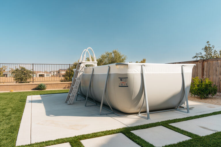 A large rectangular above-ground pool features the My Store Confer A Frame Pool Ladder on a concrete patio in a backyard, surrounded by grass, wooden and metal fencing under a clear blue sky.