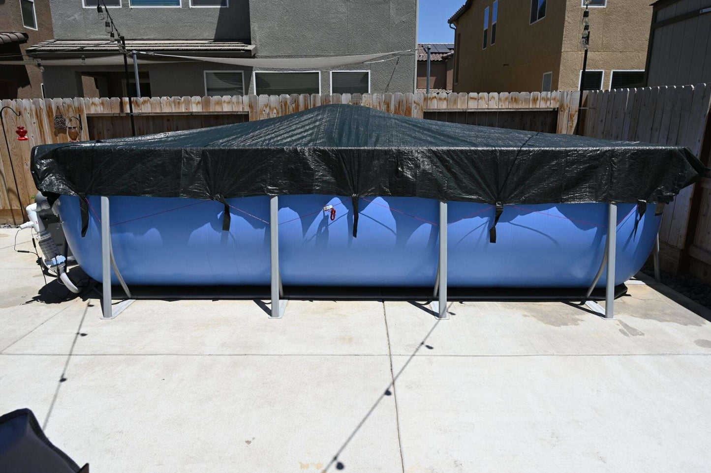 A large blue above-ground pool in a backyard is protected by a My Store Pool Cover, which is weatherproof and secured over the top, surrounded by a wooden fence and neighboring houses.