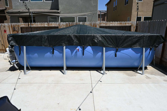 A large blue above-ground pool in a backyard is protected by a My Store Pool Cover, which is weatherproof and secured over the top, surrounded by a wooden fence and neighboring houses.