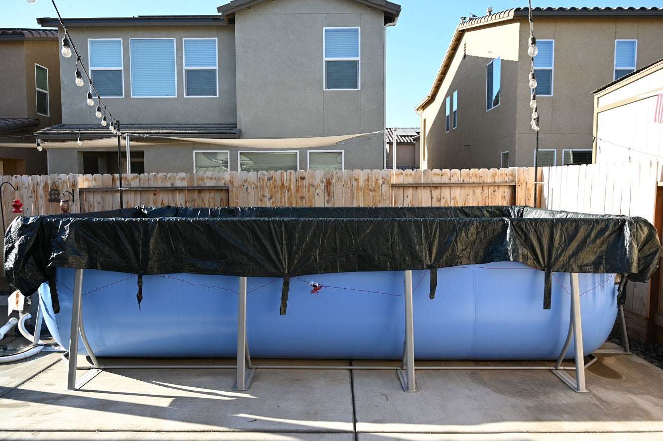 A My Store Pool Cover is partially draped over a large blue above-ground pool on a concrete patio, surrounded by a wooden fence and houses in the backyard. String lights hang overhead.