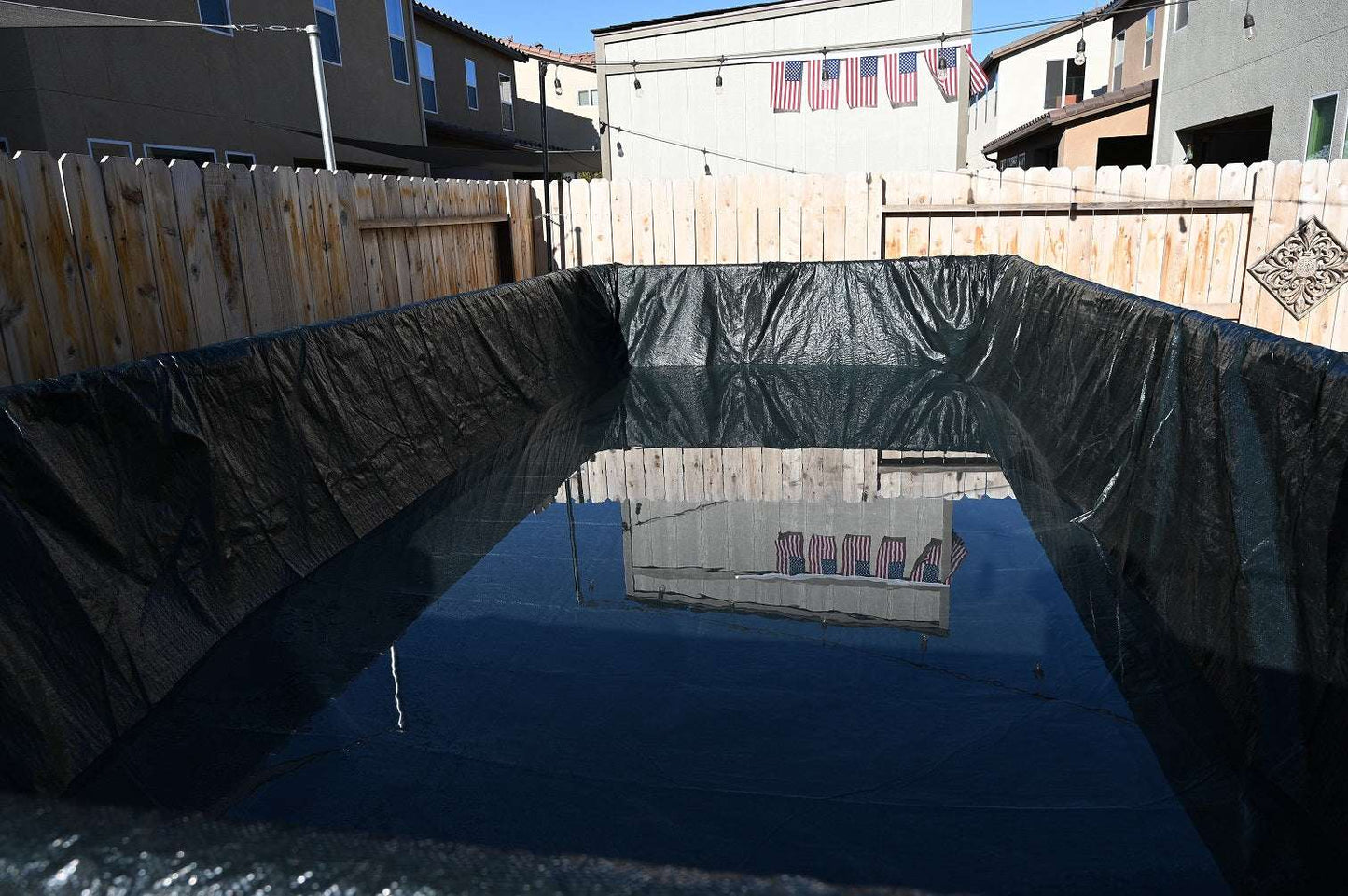 A backyard makeshift pool with a black tarp and water, enclosed by a wooden fence and nearby houses, shows the need for My Store Pool Covers for reliable pool winter protection.