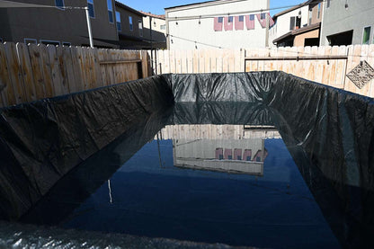 A backyard makeshift pool with a black tarp and water, enclosed by a wooden fence and nearby houses, shows the need for My Store Pool Covers for reliable pool winter protection.