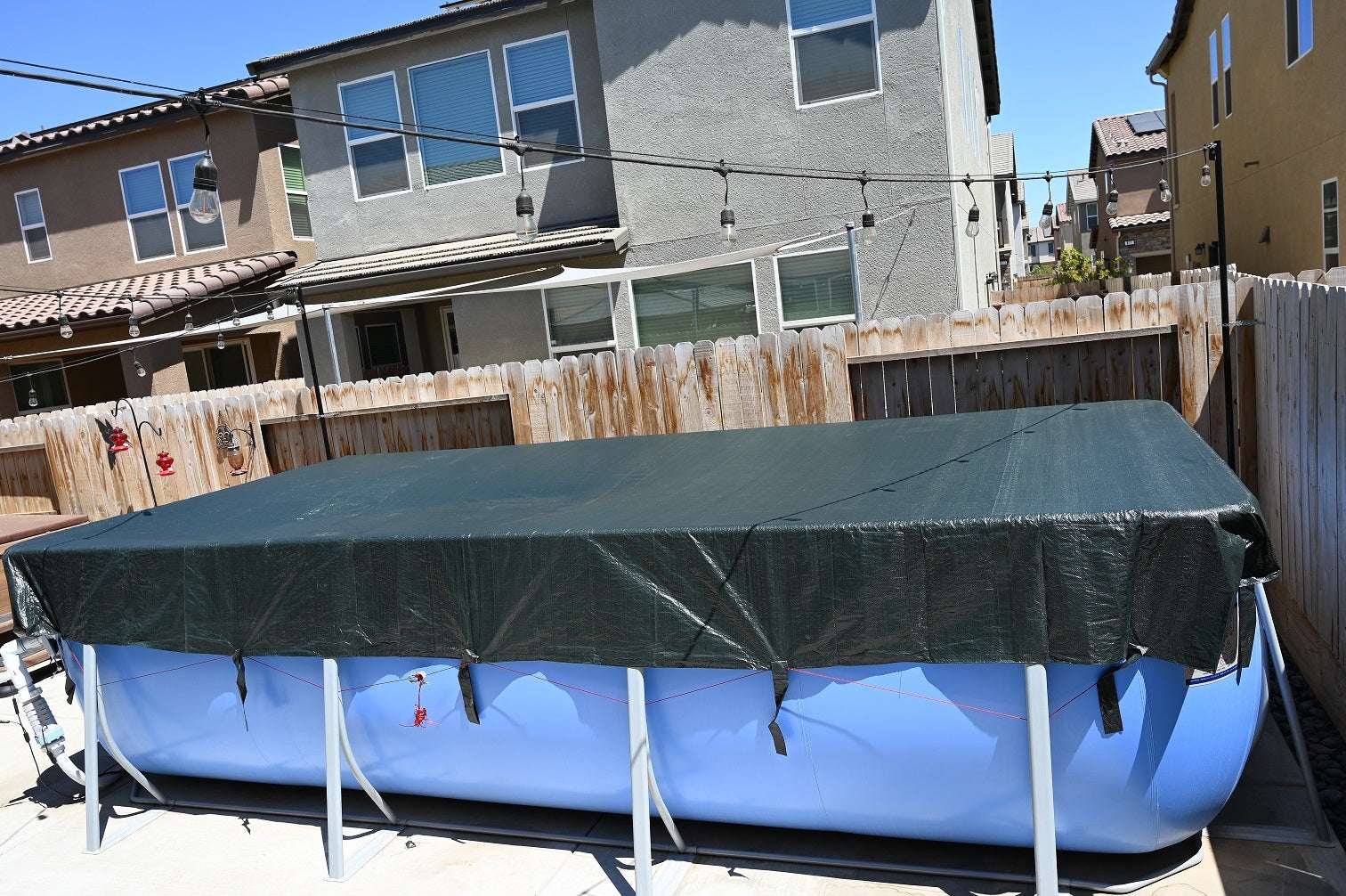 A My Store Pool Cover protects a rectangular above-ground pool in a fenced backyard. Surrounding homes and string lights are visible in the background beneath a clear blue sky.